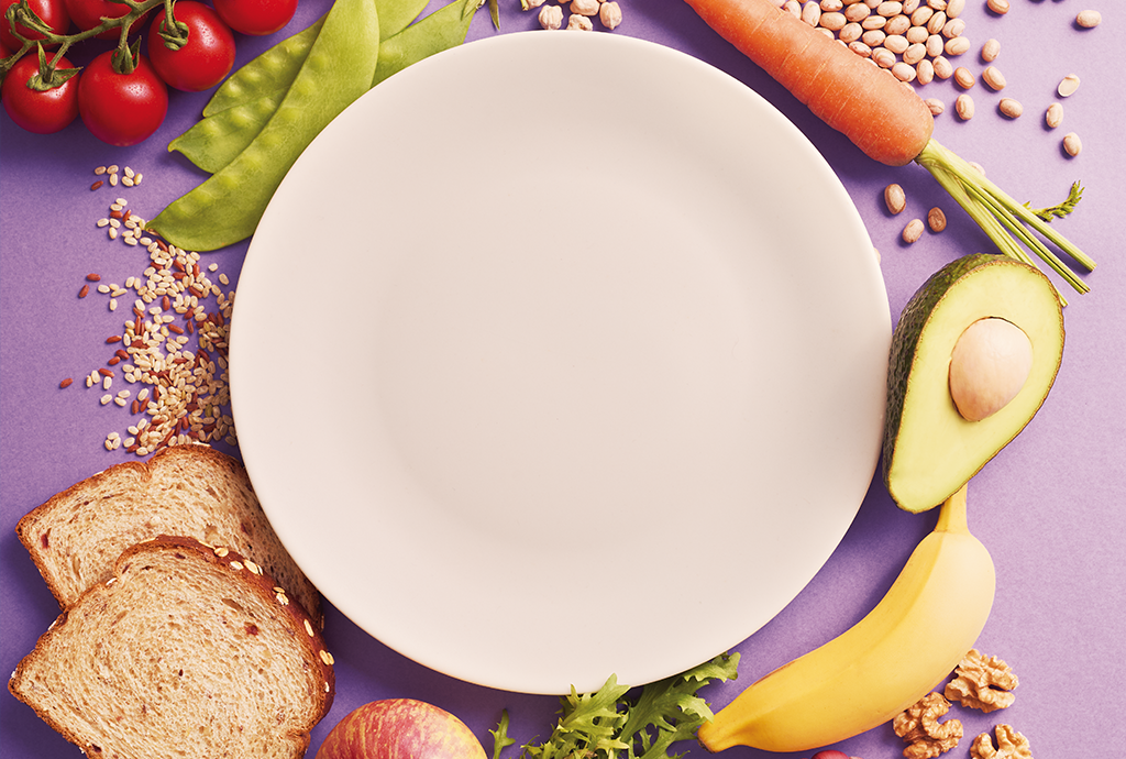 Prato branco vazio cercado por alimentos saudáveis em fundo roxo. Há tomates cereja, ervilhas, grãos, cenoura, abacate, banana, nozes, maçã, alface e pão integral