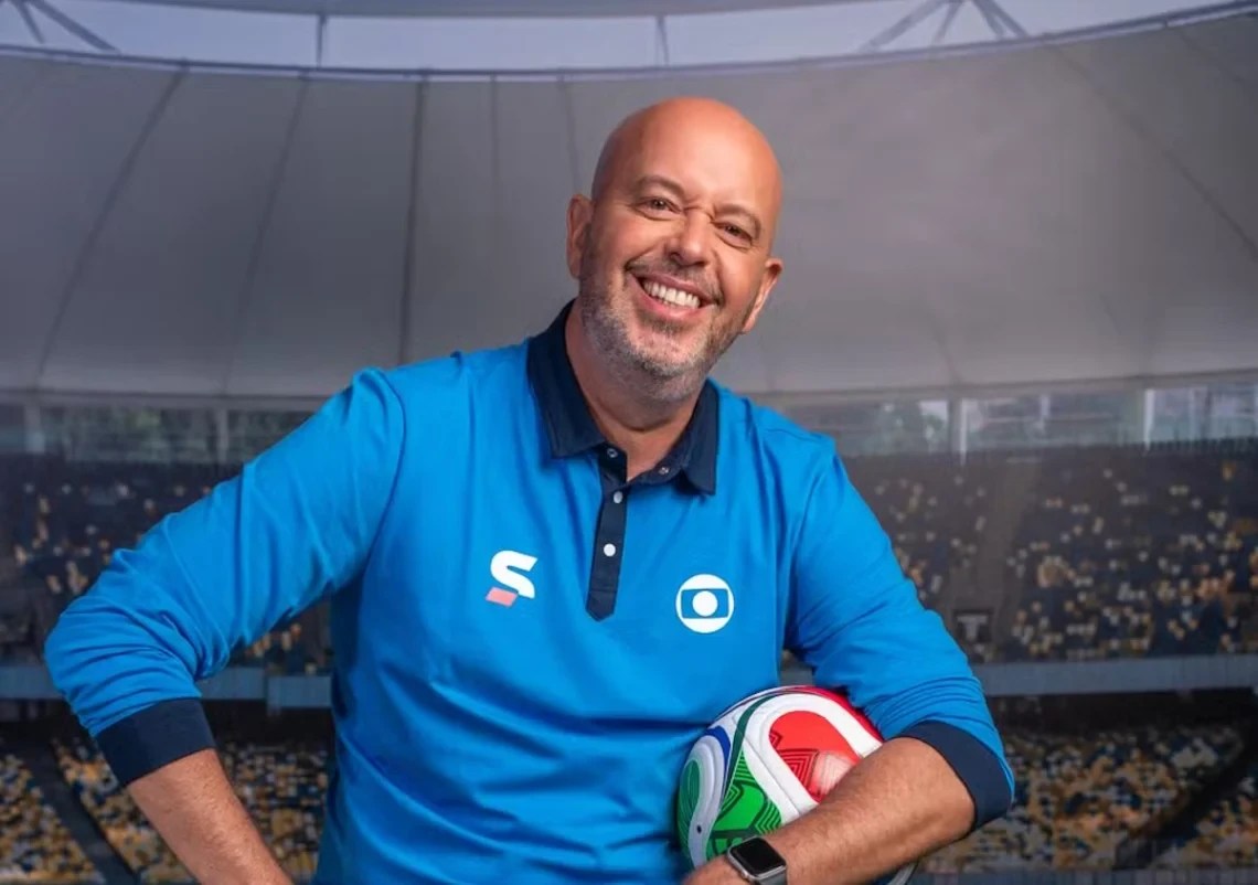 Homem careca de barba rala, vestindo camisa polo azul de manga comprida com logos S e Globo, sorri segurando uma bola de futebol colorida, em frente a um estádio com arquibancadas cheias