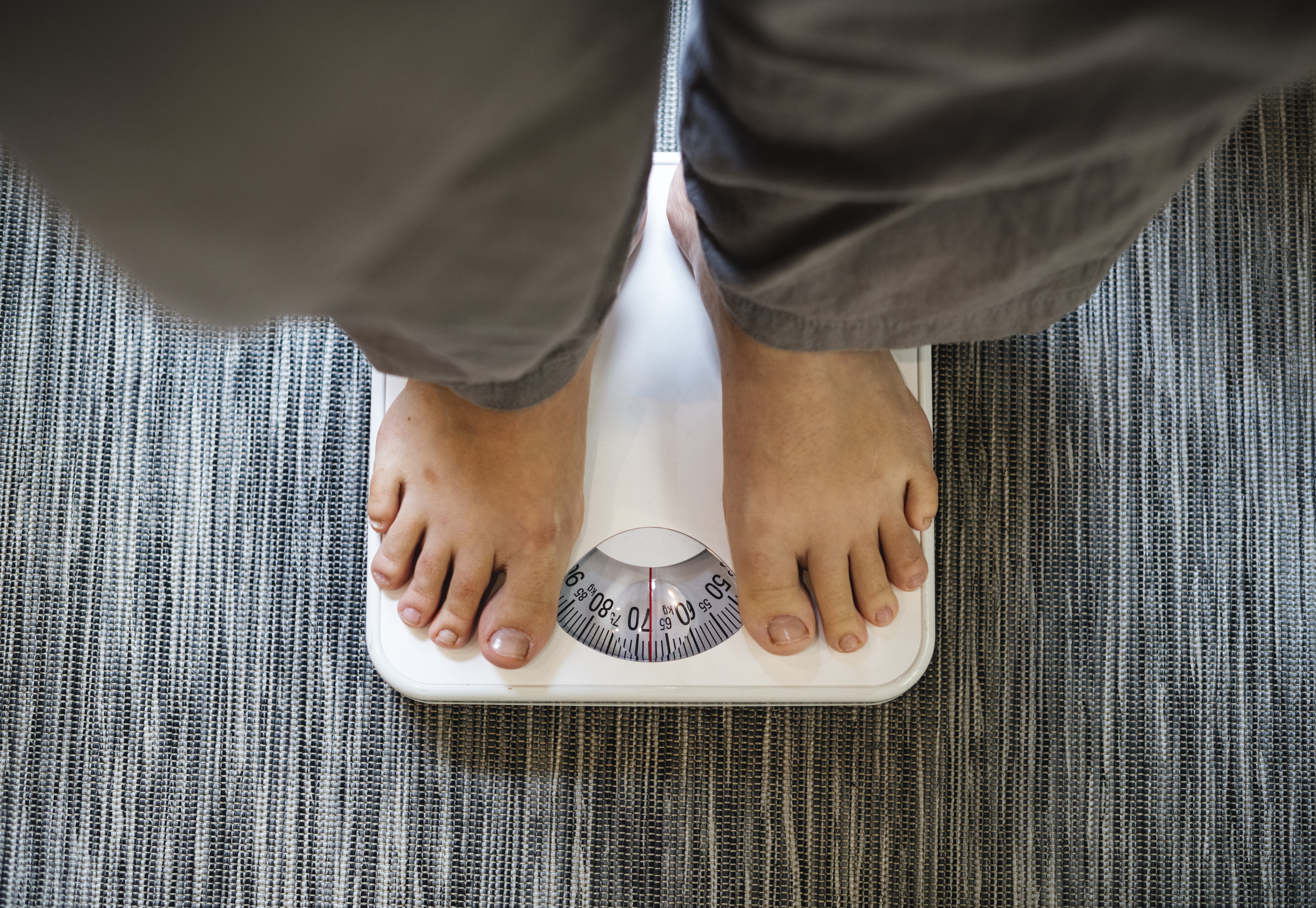 Pés de uma pessoa, vestindo calças cinzas, sobre uma balança branca analógica, que marca aproximadamente 70 kg. A balança está sobre um tapete listrado cinza e branco